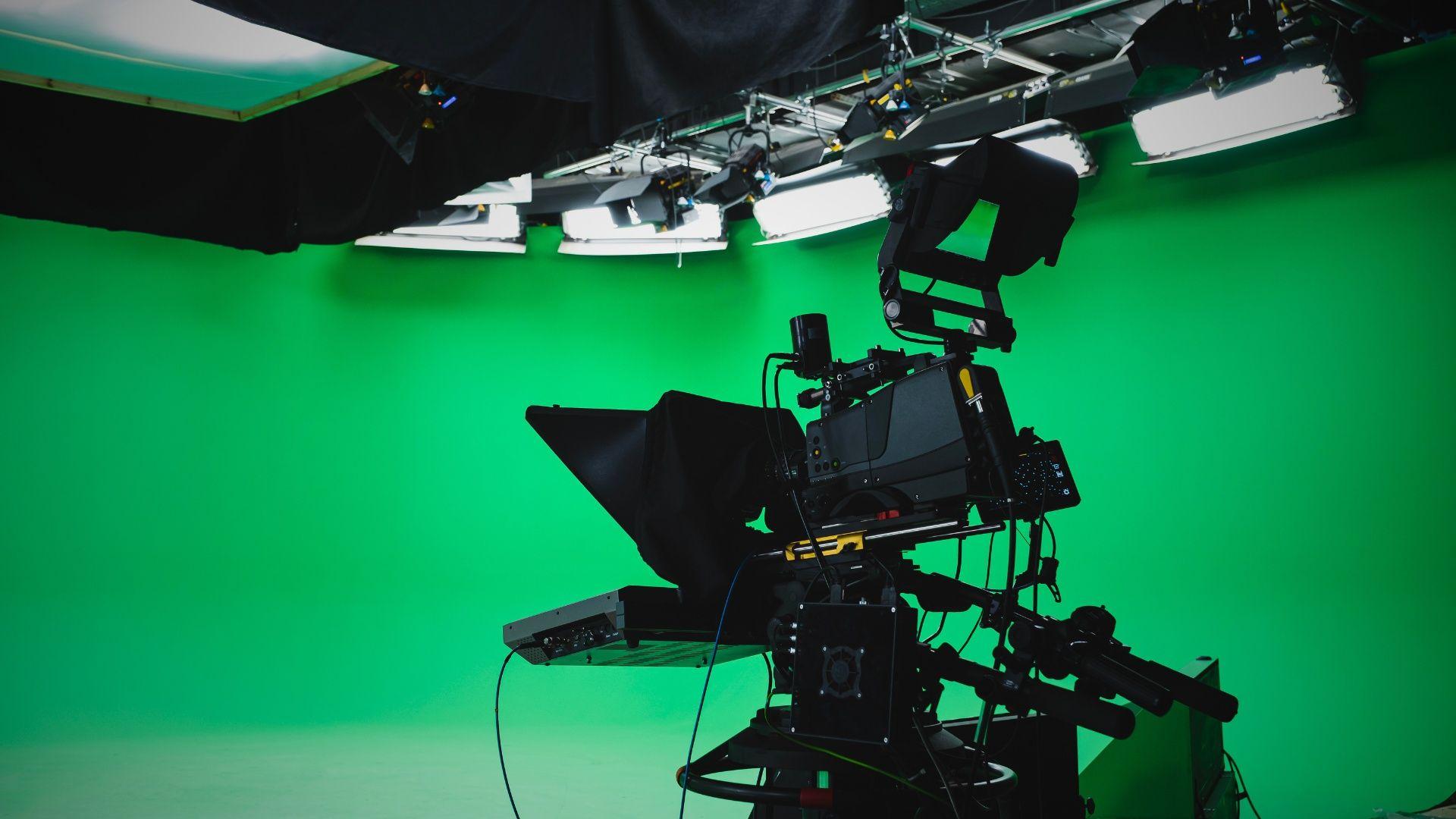 Profesjonalna kamera telewizyjna ustawiona na statywie w studiu nagraniowym z zielonym ekranem w tle, oświetlonym równomiernie przez liczne lampy sufitowe; kamera wyposażona w teleprompter, liczne przewody i akcesoria techniczne.
