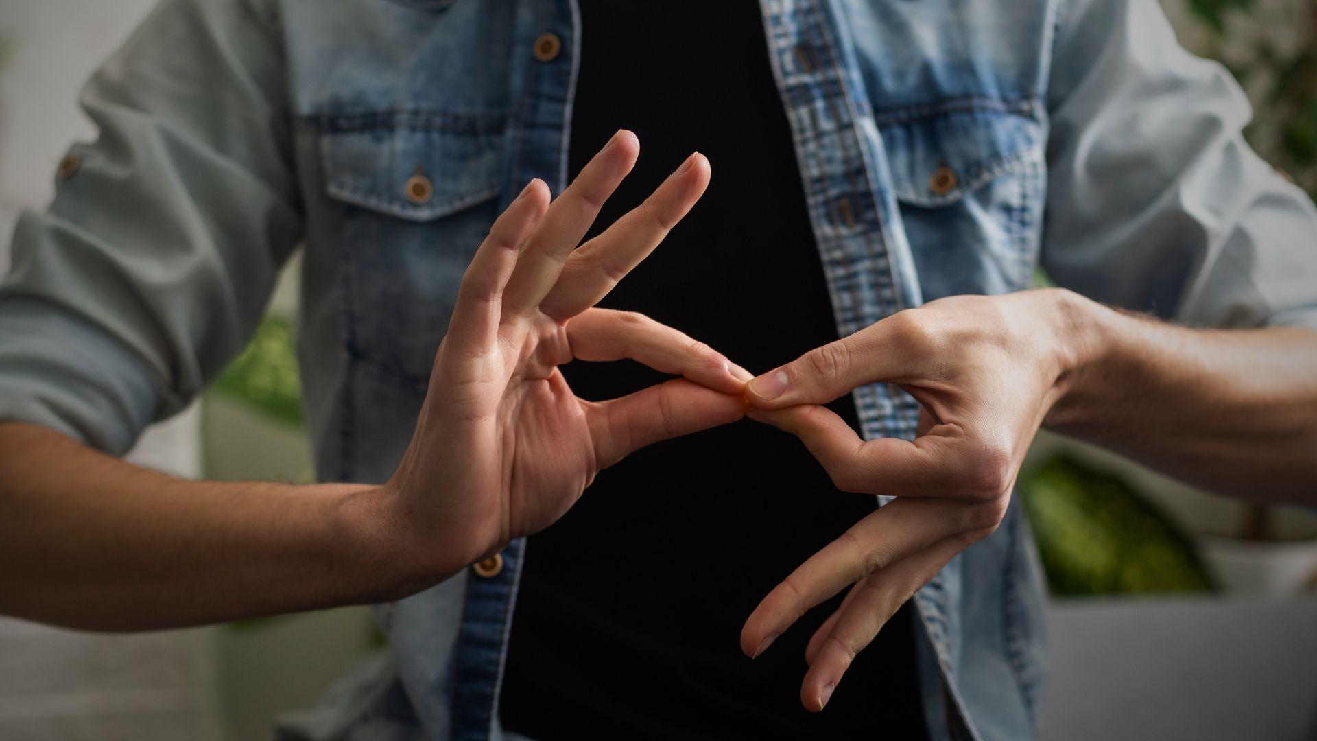 Osoba ubrana w czarną koszulkę i rozpiętą jeansową koszulę wykonuje gest języka migowego, łącząc palce obu dłoni. Tło jest rozmyte, a gest stanowi centralny punkt kadru, sugerując komunikację niewerbalną lub tematykę dostępności i integracji osób niesłyszących.